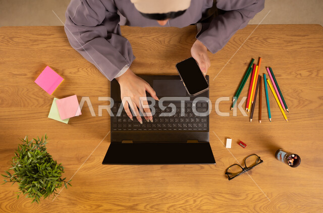 تدوين الأفكار وتسجيل المعلومات، انجاز مهام العمل عن بعد، تثبيت أوراق الملاحظات التذكارية الملونة، استخدام الأجهزة التقنية الحديثة  في الأعمال المنزلية، بورتريه علوي عامودي لإمرأة عربية خليجية عمانية تستخدم الهاتف والحاسوب بجانبها الألوان والنظارات الطبية