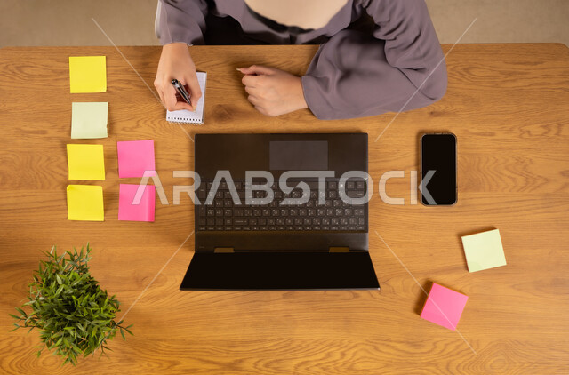 Writing down ideas and recording information, fixing colorful mementos, using modern technical devices in household chores, completing work tasks remotely, vertical upper portrait of an Arab Gulf Omani woman using a phone and laptop and writing on a notebook
