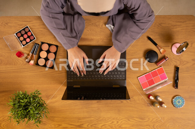 التسويق لمكياج وميكاب تتوريال، أدوات تجميل نسائية عصرية، مفهوم الأنوثة والاهتمام بالجمال والمظهر الخارجي، دمج التكنولوجيا والتقنيات الحديثة في الحياة اليومية، بورتريه علوي عامودي لإمرأة عربية خليجية عمانية تستخدم الحاسوب المحمول وبجانبها معدات تجميلية