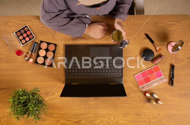 Modern women's cosmetics, the concept of femininity and interest in beauty and appearance, marketing for makeup and makeup tutorials, integrating modern technology into daily life, a vertical upper portrait of an Arab Gulf Omani woman using a laptop and holding a mirror in her hand and cosmetic equipment next to her