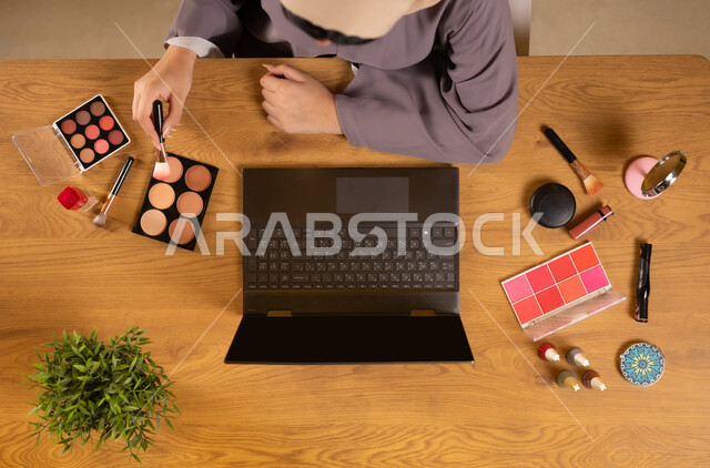 مفهوم الأنوثة والاهتمام بالجمال والمظهر الخارجي، أدوات تجميل نسائية عصرية، التسويق لمكياج وميكاب تتوريال، دمج التكنولوجيا الحديثة في الحياة اليومية، بورتريه علوي عامودي لإمرأة عربية خليجية عمانية تستخدم الحاسوب المحمول وتحمل إحدى المعدات التجميلية (الفرشاة)