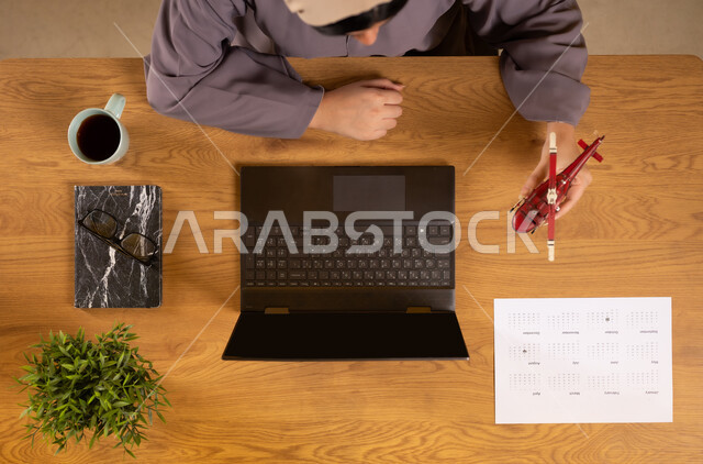 Preparing for a fun summer trip, the concept of tourism and leisure tours, vertical overhead portrait of an Omani Gulf Arab woman using a laptop and holding a model of an airplane, obtaining the necessary supplies via the Internet, integrating technology into practical life