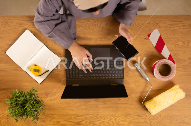 الحصول على أدوات الدهان عبر الإنترنت، الاهتمام بالتجديد والتطور، سهولة شراء مستلزمات المهن من خلال الاونلاين، استخدام التكنولوجيا والتقنيات الحديثة والمتطورة، بورتريه علوي عامودي لإمرأة عربية خليجية عمانية تستخدم الهاتف والحاسوب المحمول، عرض شاشة سوداء فارغة