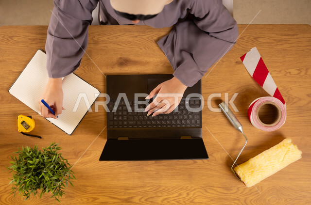 الاهتمام بالتجديد والتطور، الحصول على أدوات الدهان عبر الإنترنت، سهولة شراء مستلزمات المهن من خلال الاونلاين، استخدام التكنولوجيا والتقنيات الحديثة والمتطورة، بورتريه علوي عامودي لإمرأة عربية خليجية عمانية تستخدم الحاسوب وتقوم بتدوين الملاحظات على الدفتر