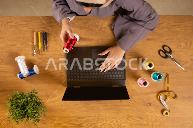 الإستفادة من المواقع الإلكترونية للتعلم والتدريب، الحصول على أدوات الخياطة أونلاين، بورتريه علوي عامودي لإمرأة عربية خليجية عمانية تستخدم الحاسوب المحمول تمسك بيدها الخيطان وبجانبها مستلزمات الحياكة، عرض شاشة سوداء فارغة، استخدام التكنولوجيا والتقنيات الحديثة