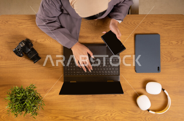 دمج التكنولوجيا في الحياة اليومية، استخدام التقنيات الحديثة والمتطورة، بورتريه علوي عامودي لإمرأة عربية خليجية عمانية تقوم باستخدام جهاز الحاسوب المحمول والهاتف بشاشة سوداء فارغة، التدريب من خلال الإنترنت، الحصول على المستلزمات اللازمة عن طريق الأونلاين