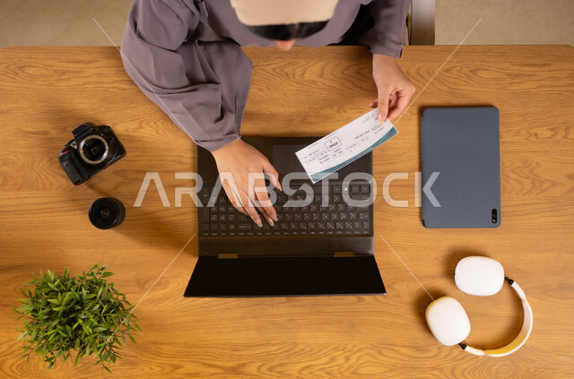 The concept of tourism and leisure tours, preparing for a fun summer trip, vertical overhead portrait of an Omani Gulf Arab woman using a laptop and holding a travel visa, obtaining the necessary supplies online, integrating technology into practical life