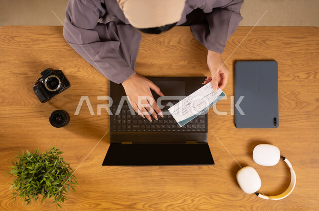 الاستعداد لقضاء رحلة صيفية ممتعة، مفهوم السياحة والقيام بالجولات الترفيهية، بورتريه علوي عامودي لإمرأة عربية خليجية عمانية تستخدم جهاز الحاسوب المحمول تحمل بيدها تأشيرة السفر، الحصول على المستلزمات اللازمة عن طريق الاونلاين، دمج التكنولوجيا في الحياة العملية