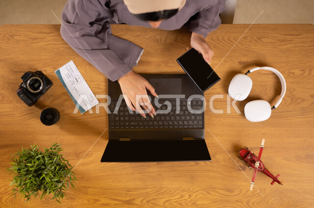 Integrating technology into practical life, preparing for a fun summer trip, the concept of tourism and leisure tours, vertical overhead portrait of an Arab Gulf Omani woman using a phone and laptop, holding a travel visa in her hand, obtaining the necessary supplies through the Internet