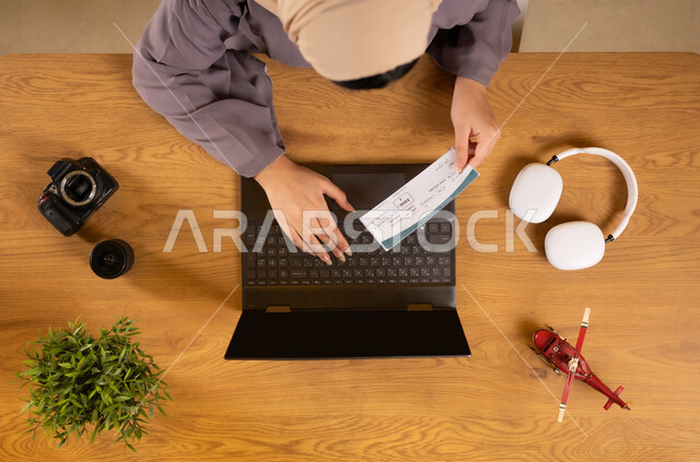 الاستعداد لقضاء رحلة صيفية ممتعة، مفهوم السياحة والقيام بالجولات الترفيهية، بورتريه علوي عامودي لإمرأة عربية خليجية عمانية تستخدم جهاز الحاسوب المحمول تحمل بيدها تأشيرة السفر، الحصول على المستلزمات اللازمة عن طريق الاونلاين، دمج التكنولوجيا في الحياة العملية