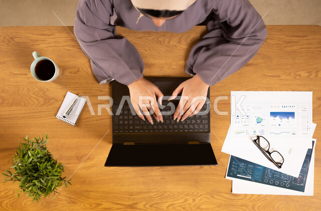 دمج الأجهزة التقنية الحديثة في الحياة العملية، متابعة مخطط الرسوم البيانية، الإستثمار المالي الإلكتروني، مراقبة الإقتصاد والمعلومات المالية، بورتريه علوي عامودي لإمرأة عربية خليجية عمانية تقوم بدراسة وتحليل الإحصائيات عبر جهاز الحاسوب المحمول