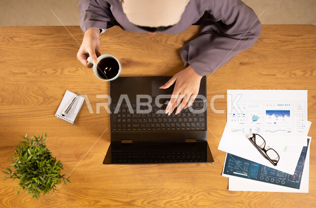 الإستثمار المالي الإلكتروني، دمج الأجهزة التقنية الحديثة في الحياة العملية، متابعة مخطط الرسوم البيانية، مراقبة الإقتصاد والمعلومات المالية، بورتريه علوي عامودي لإمرأة عربية خليجية عمانية تقوم بدراسة وتحليل الإحصائيات عبر جهاز الحاسوب المحمول وترتشف القهوة