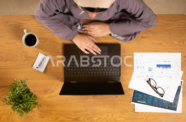 دمج الأجهزة التقنية الحديثة في الحياة العملية، متابعة مخطط الرسوم البيانية، الإستثمار المالي الإلكتروني، مراقبة الإقتصاد والمعلومات المالية، بورتريه علوي عامودي لإمرأة عربية خليجية عمانية تقوم بدراسة وتحليل الإحصائيات عبر جهاز الحاسوب المحمول