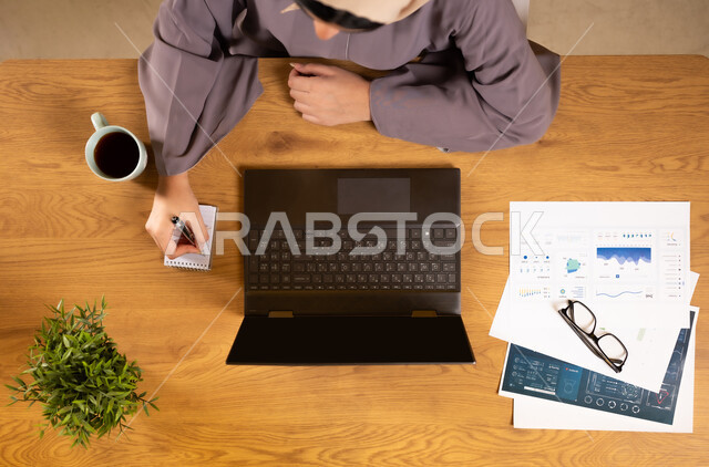 متابعة مخطط الرسوم البيانية، تدوين الملاحظات على الدفتر، مراقبة الإقتصاد والمعلومات المالية، دمج الأجهزة التقنية الحديثة بالحياة العملية، الإستثمار المالي الإلكتروني، بورتريه علوي عامودي لإمرأة عربية خليجية عمانية تقوم بدراسة وتحليل الإحصائيات عبر جهاز الحاسوب