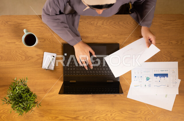 Integrating modern technology devices into practical life, following up on charts, electronic financial investment, monitoring the economy and financial information, vertical top portrait of an Arab Gulf Omani woman studying and analyzing statistics via a laptop