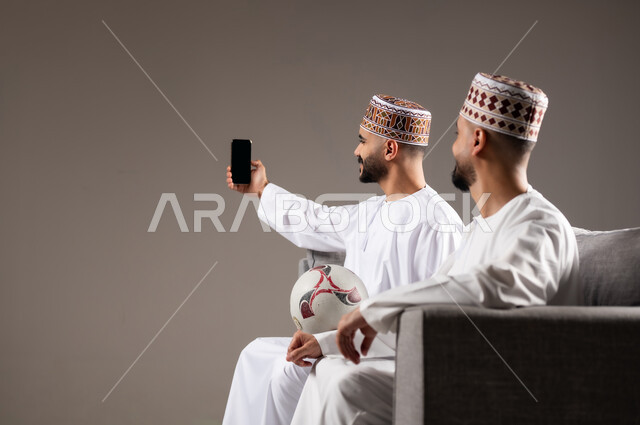 Taking souvenir photos, spending quality time with family and friends, gestures of joy, pleasure and happiness, portrait of two Omani Gulf Arab men wearing dishdasha and thobe, one of them holding a mobile phone and taking a picture, gray background