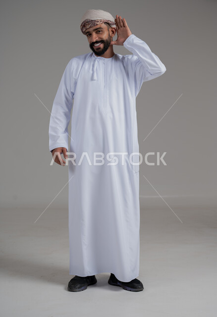 ايماءات وجه ويدين تدل على السعادة والسرور، وضع اليد خلف الاذن لرصد الصوت القادم، بورتريه لرجل عربي خليجي عماني يرتدي الدشداشه والعمامة يضع يده وراء اذنه لمحاولة السمع، الجسم بالطول الكامل، خلفية بيضاء 