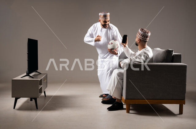 التقاط الصور التذكارية, قضاء وقت ممتع مع العائلة والاصدقاء, ايماءات الفرح والسرور والسعادة, بورتريه لرجلان عربيان عمانين خليجيان يرتديان الدشداشة والكمة يمسك احدهم هاتف محمول ويلتقط صورة, خلفية رمادية