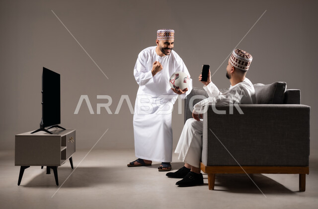 Taking souvenir photos, spending quality time with family and friends, gestures of joy, pleasure and happiness, portrait of two Omani Gulf Arab men wearing dishdasha and thobe, one of them holding a mobile phone and taking a picture, gray background