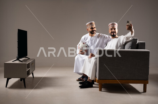 التقاط الصور التذكارية, قضاء وقت ممتع مع العائلة والاصدقاء, ايماءات الفرح والسرور والسعادة, بورتريه لرجلان عربيان عمانين خليجيان يرتديان الدشداشة والكمة يمسك احدهم هاتف محمول ويلتقط صورة, خلفية رمادية