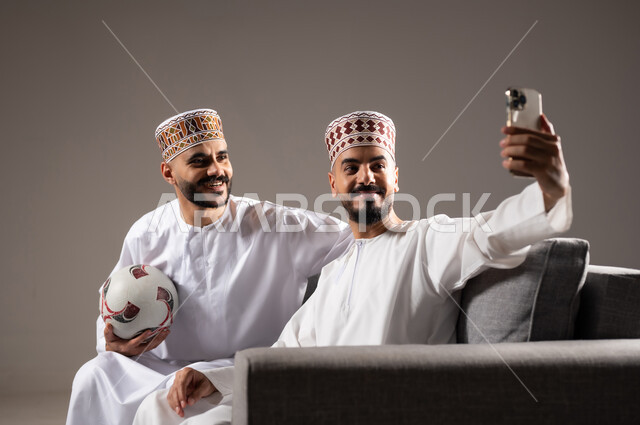 Taking souvenir photos, spending quality time with family and friends, gestures of joy, pleasure and happiness, portrait of two Omani Gulf Arab men wearing dishdasha and thobe, one of them holding a mobile phone and taking a picture, gray background