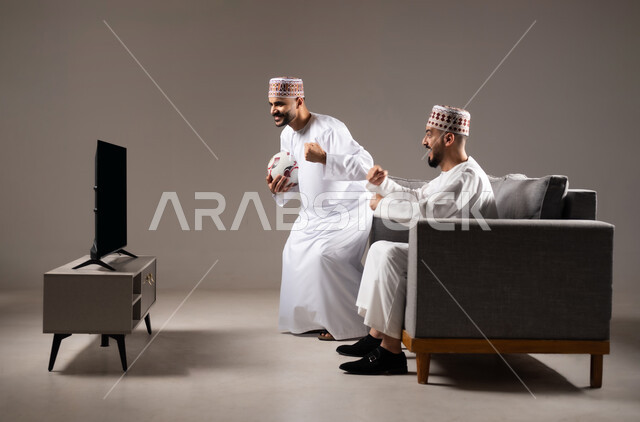 Interaction and integration by watching the favorite team, matches and sports games, portrait of two Omani Gulf Arab men wearing the dishdasha and the kameh, one of them standing with gestures of enthusiasm, victory and triumph, passion for recreational activities, gray background