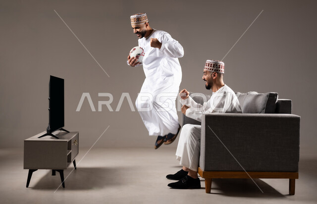 التفاعل والاندماج بمشاهدة الفريق المفضل, المباريات والالعاب الرياضية, بورتريه لرجلان عربيان عمانين خليجيان يرتديان الدشداشة والكمة احدهما يقف بإيماءات الحماس و الفوز والانتصار، الشغف بالانشطة الترفيهية, خلفية رمادية