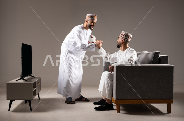 Interaction and integration by watching the favorite team, matches and sports games, portrait of two Omani Gulf Arab men wearing the dishdasha and the kameh, one of them standing with gestures of enthusiasm, victory and triumph, passion for recreational activities, gray background