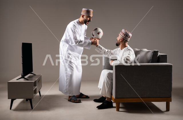 Interaction and integration by watching the favorite team, matches and sports games, portrait of two Omani Gulf Arab men wearing the dishdasha and the kameh, one of them standing with gestures of enthusiasm, victory and triumph, passion for recreational activities, gray background