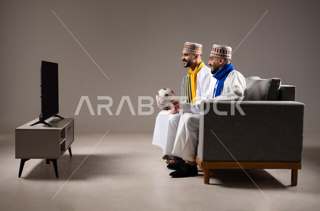 المباريات والالعاب الرياضية, التفاعل والاندماج بمشاهدة الفريق المفضل, ايماءات الفرح والحماس بالفوز والانتصار, بورتريه لرجلان عربيان عمانين خليجيان يرتديان الدشداشة والكمة احدهما يمسك بيده كرة قدم, الشغف بالانشطة الترفيهية, خلفية رمادية