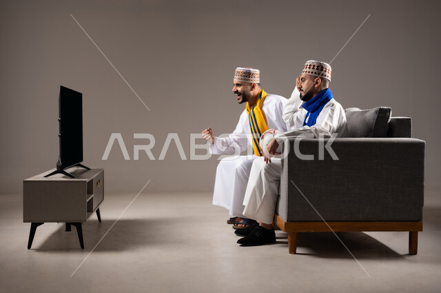 Sports matches and games, interaction and integration by watching the favorite team, gestures of joy and enthusiasm for winning and triumph, portrait of two Arab Omani Gulf men wearing the dishdasha and the kummah, one of them holding a football in his hand, passion for recreational activities, gray background
