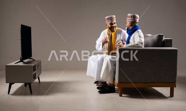 الاستمتاع بتبادل الاحاديث مع الاصدقاء, مفهوم العلاقة القوية والترابط بين الزملاء, تعابير التسلية والمرح, بورتريه لرجلان عربيان عمانيان خليجيان يرتدون الدشداشة مع الكمه ويجلسون على الاريكه, نشاط منزلي ترفيهي جماعي, قضاء اوقات ممتعة ومسلية, خلفية رمادية