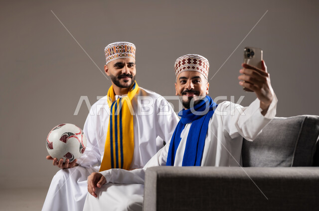 قضاء وقت ممتع مع العائلة والاصدقاء, التقاط الصور التذكارية, ايماءات الفرح والسرور والسعادة, بورتريه لرجلان عربيان عمانين خليجيان يرتديان الدشداشة والكمة يمسك احدهم هاتف محمول ويلتقط صورة, خلفية رمادية