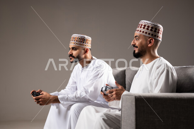 قضاء وقت ممتع, المتعة و التسلية والتفاعل بالبلايستيشن الترفيهية,  بورتريه لرجلان عربيان خليجيان عمانيان يجلسون في غرفة المعيشة على الأريكه ويمسكون بيديهم عصا التحكم, لعب العاب الفيديو بإيماءات تدل على السعادة والسرور، خلفية رمادية