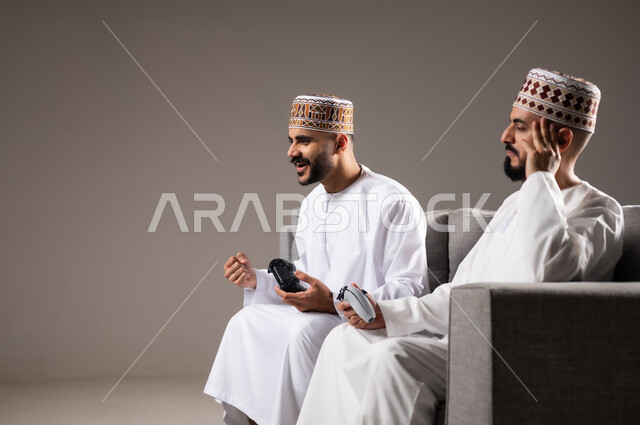المتعة و التسلية والتفاعل بالبلايستيشن الترفيهية, قضاء وقت ممتع, بورتريه لرجلان عربيان خليجيان عمانيان يجلسون في غرفة المعيشة على الأريكه ويمسكون بيديهم عصا التحكم, لعب العاب الفيديو بإيماءات تدل على السعادة والسرور، خلفية رمادية