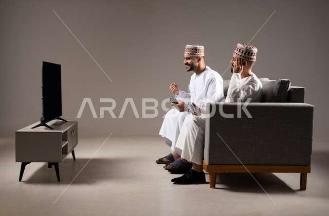 Having a good time, having fun and interacting with the PlayStation entertainment, portrait of two Omani Gulf Arab men sitting in the living room on the sofa and holding the joystick in their hands, playing video games with gestures indicating happiness and pleasure, gray background