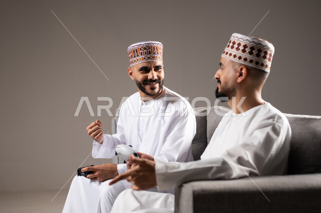 المتعة و التسلية والتفاعل بالبلايستيشن الترفيهية, قضاء وقت ممتع, بورتريه لرجلان عربيان خليجيان عمانيان يجلسون في غرفة المعيشة على الأريكه ويمسكون بيديهم عصا التحكم, لعب العاب الفيديو بإيماءات تدل على السعادة والسرور، خلفية رمادية