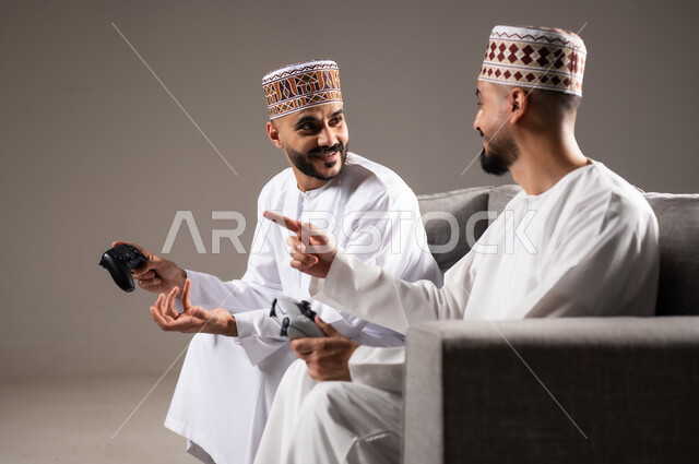 المتعة و التسلية والتفاعل بالبلايستيشن الترفيهية, قضاء وقت ممتع, بورتريه لرجلان عربيان خليجيان عمانيان يجلسون في غرفة المعيشة على الأريكه ويمسكون بيديهم عصا التحكم, لعب العاب الفيديو بإيماءات تدل على السعادة والسرور، خلفية رمادية