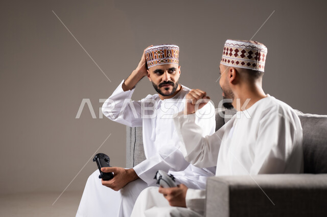 المتعة و التسلية والتفاعل بالبلايستيشن الترفيهية, قضاء وقت ممتع, بورتريه لرجلان عربيان خليجيان عمانيان يجلسون في غرفة المعيشة على الأريكه ويمسكون بيديهم عصا التحكم, لعب العاب الفيديو بإيماءات تدل على السعادة والسرور، خلفية رمادية