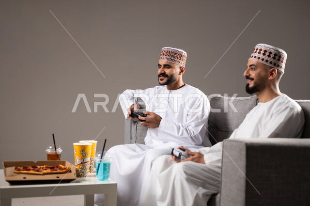 تناول الوجبات السريعة والمشاريب المنعشة, التفاعل والاندماج بمشاهدة الفريق المفضل, المباريات والالعاب الرياضية، بورتريه لرجلان عربيان عمانين خليجيان يرتديان الدشداشة والكمة احدهما يمسك بيده كرة قدم, الشغف بالانشطة الترفيهية, خلفية رمادية