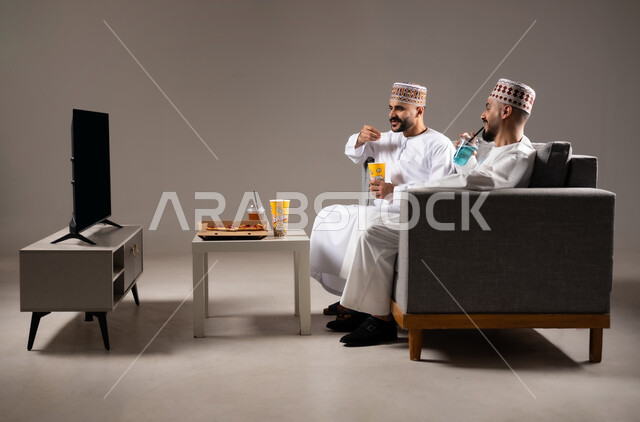 تناول الوجبات السريعة والمشاريب المنعشة, التفاعل والاندماج بمشاهدة الفريق المفضل, المباريات والالعاب الرياضية، بورتريه لرجلان عربيان عمانين خليجيان يرتديان الدشداشة والكمة احدهما يمسك بيده كرة قدم, الشغف بالانشطة الترفيهية, خلفية رمادية