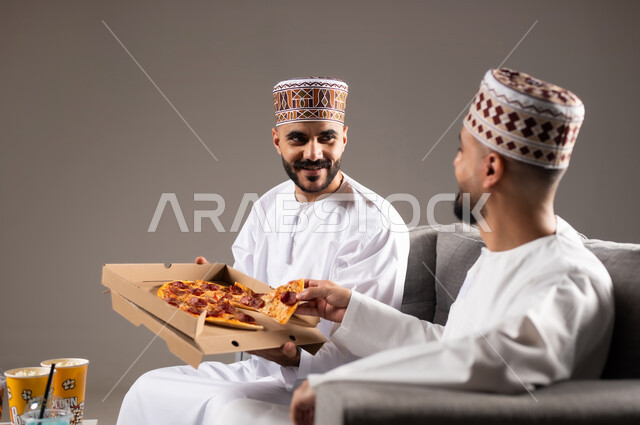 المباريات والالعاب الرياضية, تناول الوجبات السريعة والمشاريب المنعشة, بورتريه لرجلان عربيان عمانين خليجيان يرتديان الدشداشة والكمة احدهما يمسك قطعة من بيتزا, الشغف بالانشطة الترفيهية, خلفية رمادية