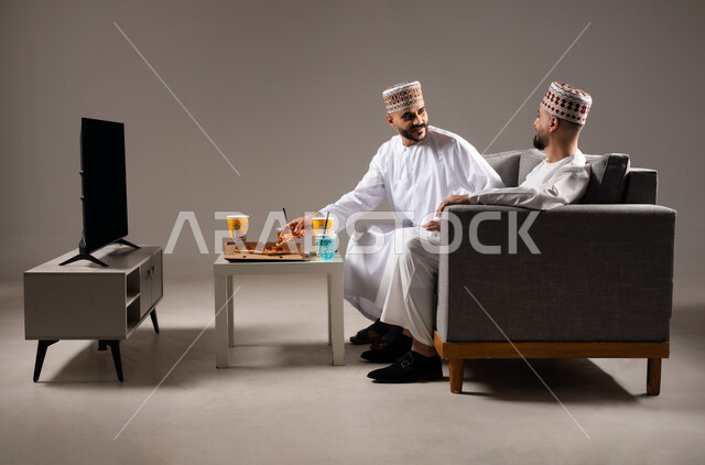 المباريات والالعاب الرياضية, تناول الوجبات السريعة والمشاريب المنعشة, بورتريه لرجلان عربيان عمانين خليجيان يرتديان الدشداشة والكمة احدهما يمسك قطعة من بيتزا, الشغف بالانشطة الترفيهية, خلفية رمادية