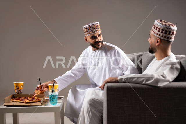 المباريات والالعاب الرياضية, تناول الوجبات السريعة والمشاريب المنعشة, بورتريه لرجلان عربيان عمانين خليجيان يرتديان الدشداشة والكمة احدهما يمسك قطعة من بيتزا, الشغف بالانشطة الترفيهية, خلفية رمادية