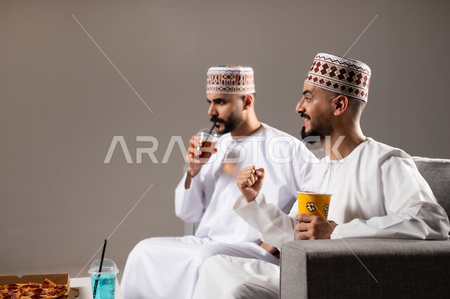 المباريات والالعاب الرياضية, التفاعل والاندماج بمشاهدة الفريق المفضل, تناول الوجبات السريعة والمشاريب المنعشة, بورتريه لرجلان عربيان عمانين خليجيان يرتديان الدشداشة والكمة يشاهدان مباراة كرة قدم، الشغف بالانشطة الترفيهية, خلفية رمادية