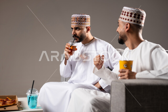 المباريات والالعاب الرياضية, التفاعل والاندماج بمشاهدة الفريق المفضل, تناول الوجبات السريعة والمشاريب المنعشة, بورتريه لرجلان عربيان عمانين خليجيان يرتديان الدشداشة والكمة يشاهدان مباراة كرة قدم، الشغف بالانشطة الترفيهية, خلفية رمادية