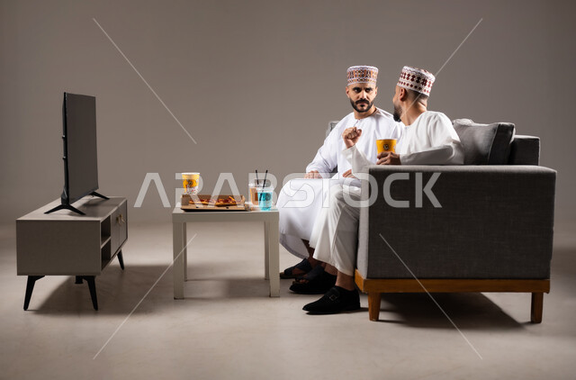 Matches and sports games, eating fast food and refreshing drinks, interaction and integration by watching the favorite team, gestures of joy and enthusiasm for victory and victory, portrait of two Omani Gulf Arab men wearing a dishdasha and a kabbah, passion for recreational activities, gray background