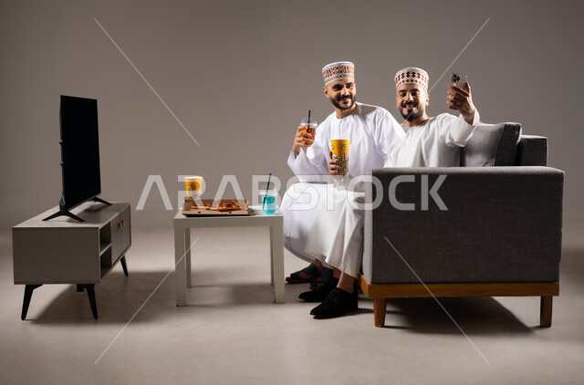 التقاط الصور التذكارية, تناول الوجبات السريعة والمشاريب المنعشة, قضاء وقت ممتع مع العائلة والاصدقاء, ايماءات الفرح والسرور والسعادة, بورتريه لرجلان عربيان عمانين خليجيان يرتديان الدشداشة والكمة يمسك احدهم هاتف محمول ويلتقط صورة, خلفية رمادية