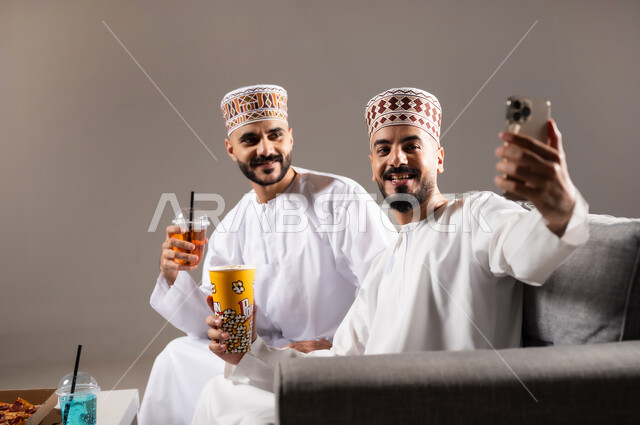 التقاط الصور التذكارية, تناول الوجبات السريعة والمشاريب المنعشة, قضاء وقت ممتع مع العائلة والاصدقاء, ايماءات الفرح والسرور والسعادة, بورتريه لرجلان عربيان عمانين خليجيان يرتديان الدشداشة والكمة يمسك احدهم هاتف محمول ويلتقط صورة, خلفية رمادية