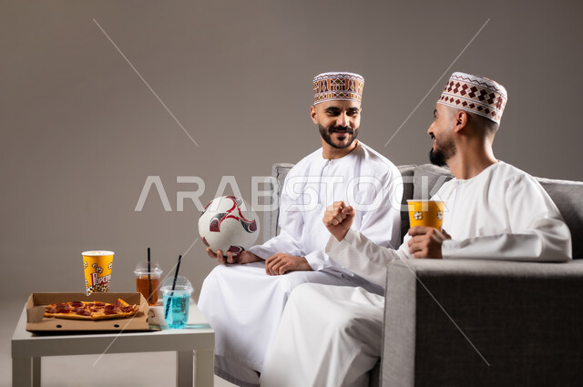 التفاعل والاندماج بمشاهدة الفريق المفضل, تناول الوجبات السريعة والمشاريب المنعشة, المباريات والالعاب الرياضية, ايماءات الفرح والحماس بالفوز والانتصار, بورتريه لرجلان عربيان عمانين خليجيان يرتديان الدشداشة والكمة احدهما يمسك بيده كرة قدم, الشغف بالانشطة الترفيهية, خلفية رمادية